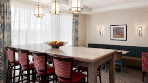A dining area with a long table, multiple chairs, hanging lights, a fruit bowl, and a wall with a framed picture.