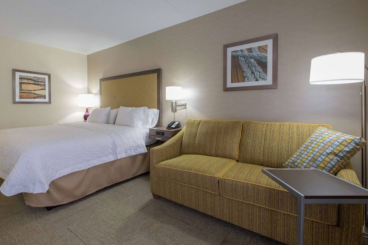 A hotel room with a bed, a sofa, a nightstand with a lamp, and a framed picture on the wall.