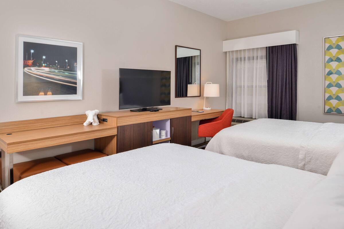 A hotel room with two beds, a desk with a TV, a chair, a lamp, and a painting on the wall.
