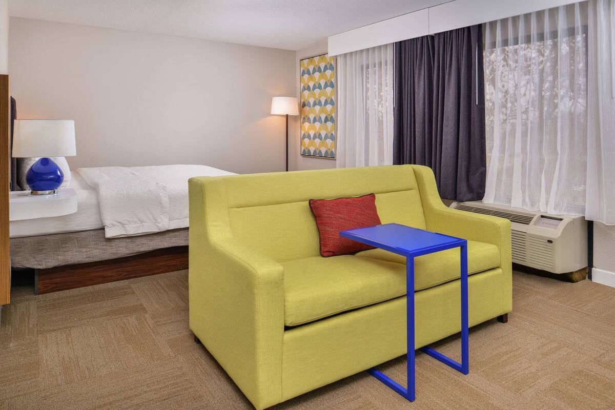 A hotel room with a yellow sofa, a small blue table, a white bed, a lamp, a wall art piece, and a window with curtains.