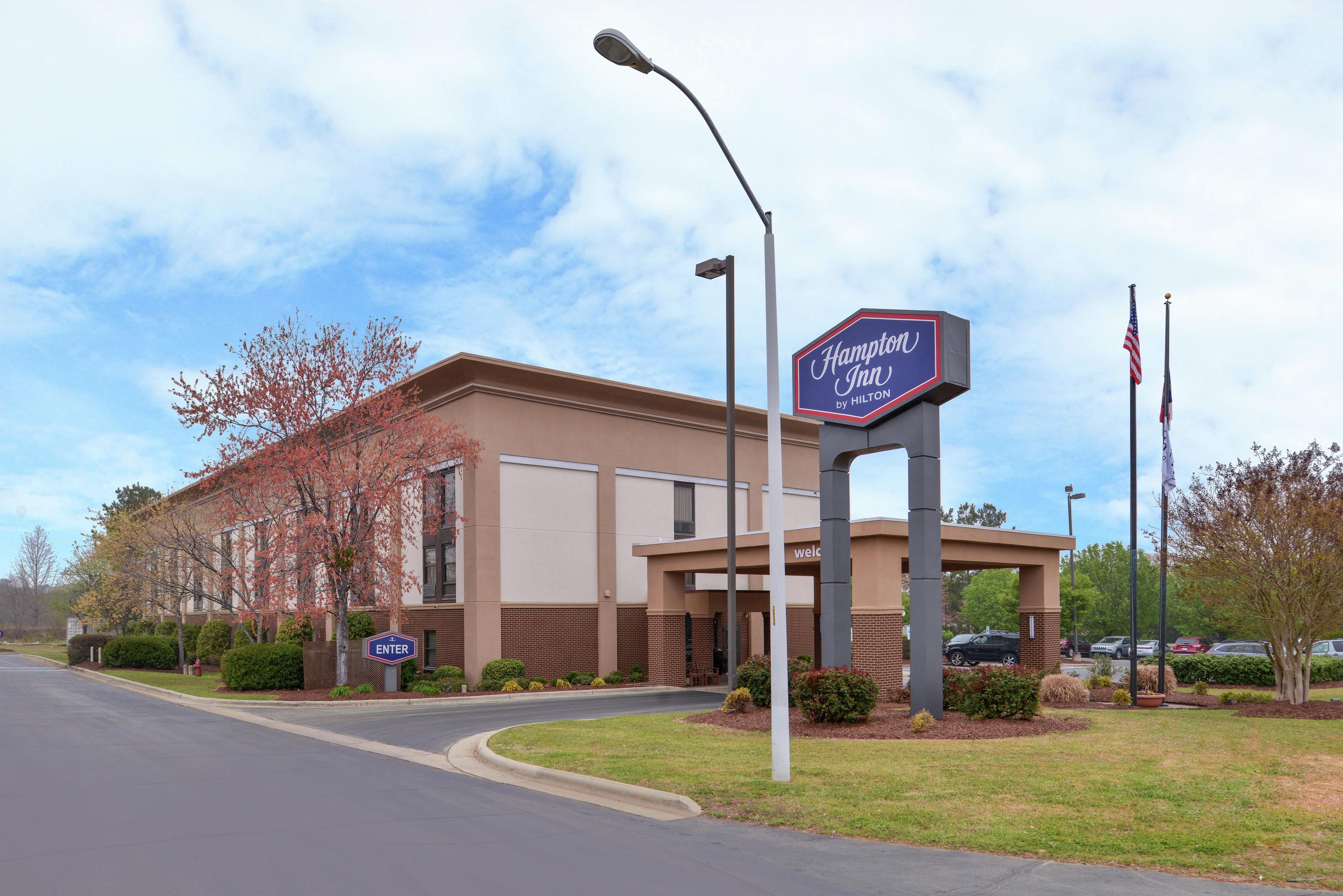 Ein Hampton Inn by Hilton Hotel mit blauem Himmel, einladendem Eingangsbereich und Parkplatz.