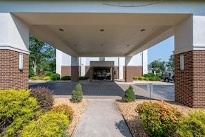 A building entrance with a covered walkway, brick walls, and a parking area with vehicles.