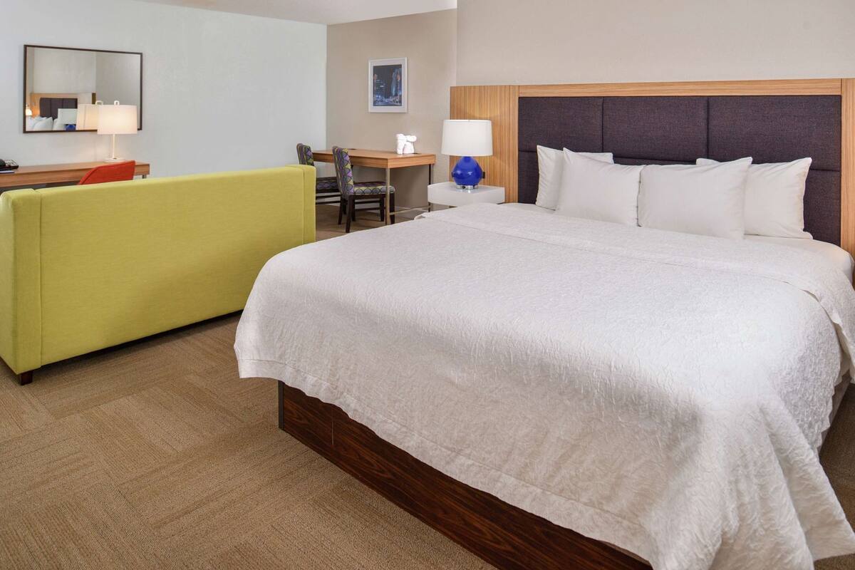 Ein Hotelzimmer mit einem großen Bett, einem gelben Sofa, einem Schreibtisch mit Lampe und einem Spiegel.