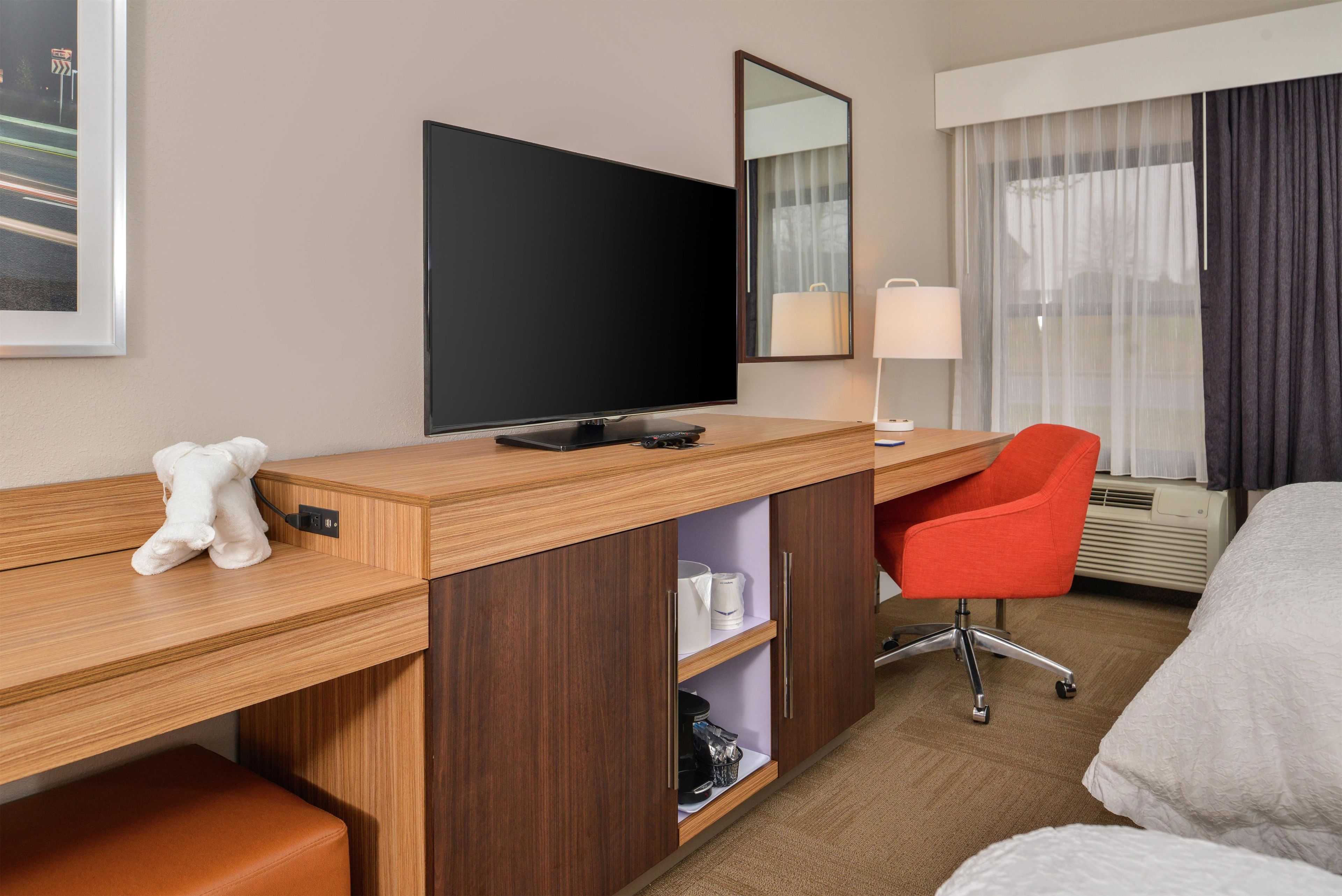 A hotel room with a wooden desk, a television, a red chair, a bed, and a window with curtains.