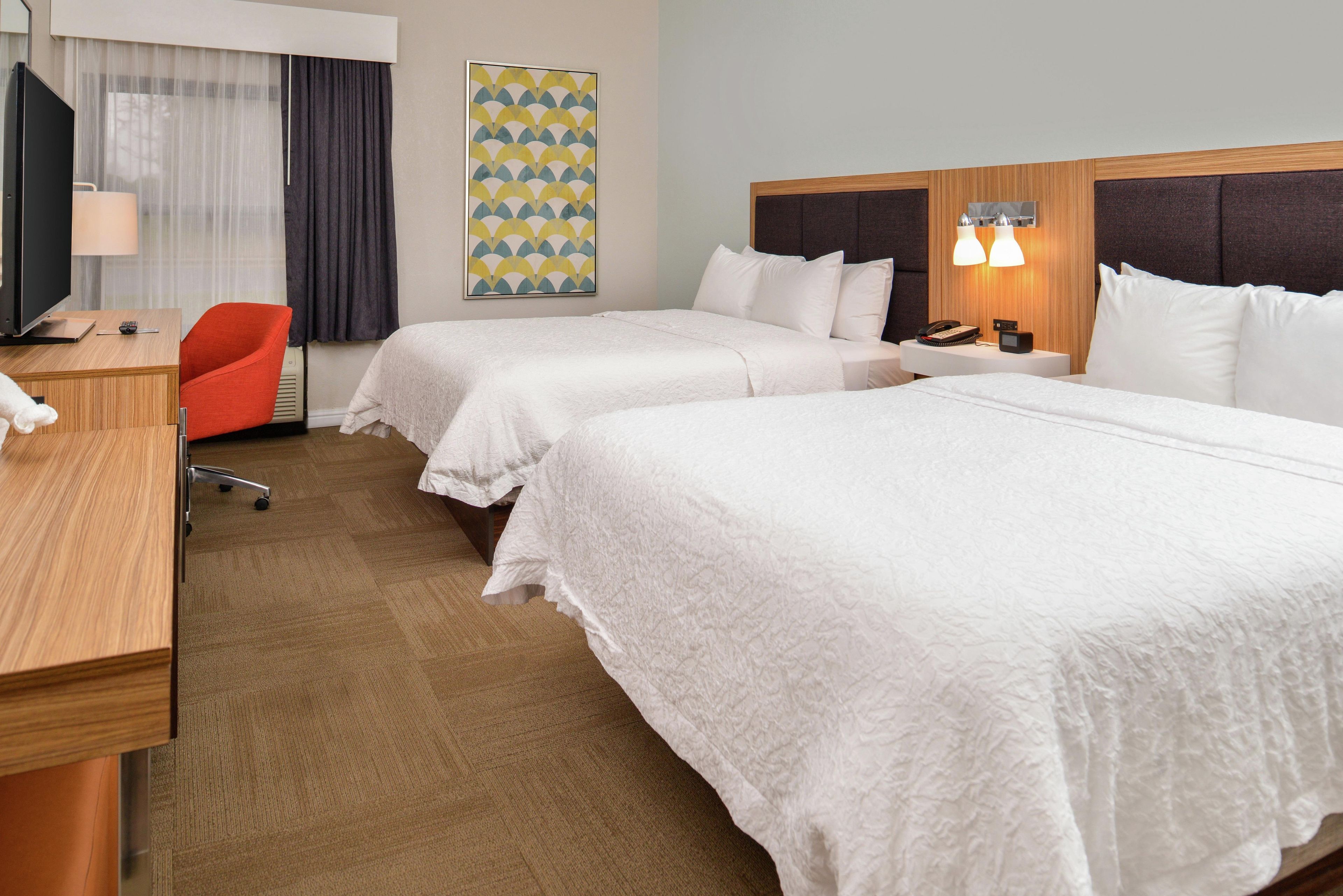 A hotel room with two beds, a desk, a chair, a TV, and a painting on the wall.