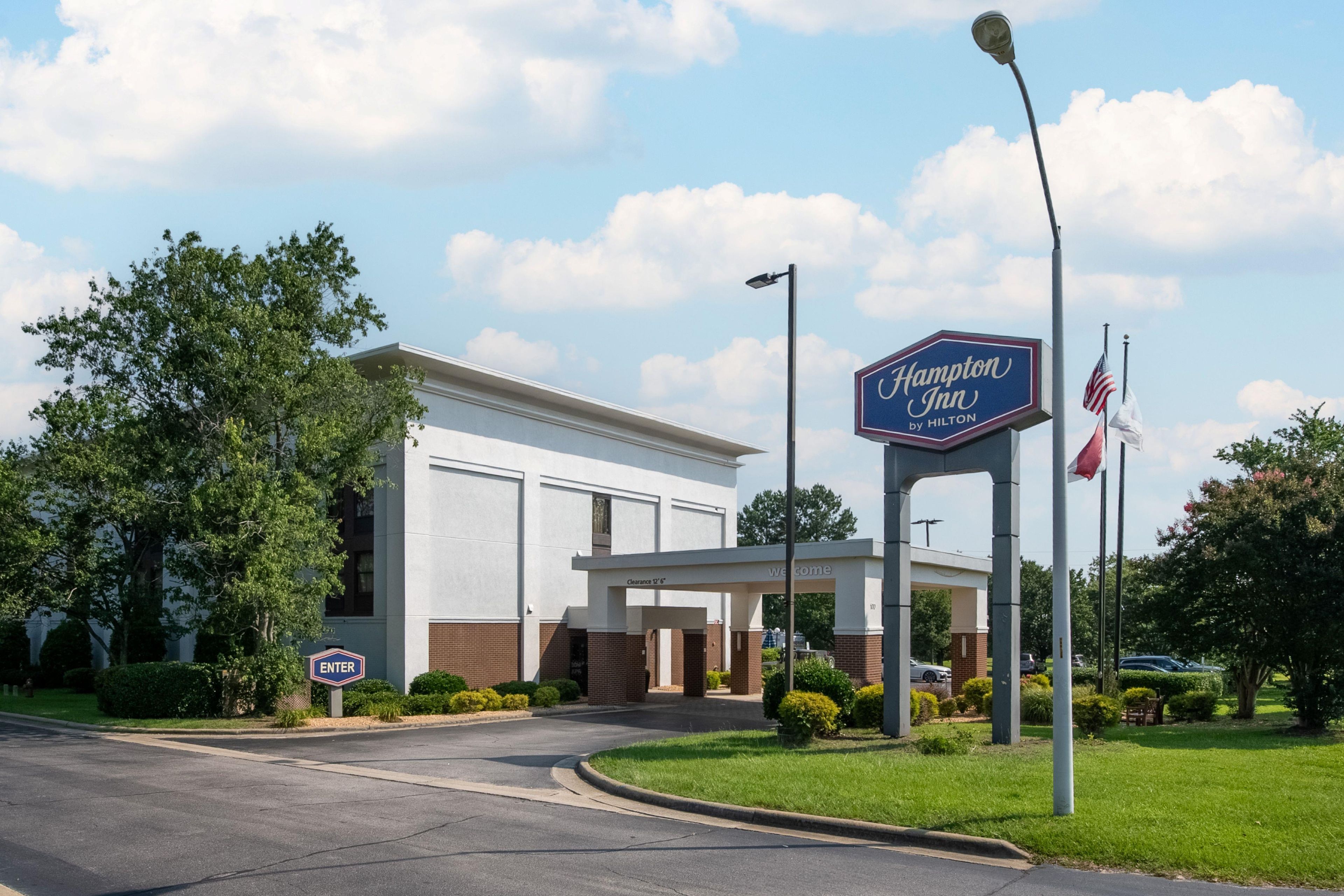 A Hampton Inn by Hilton hotel entrance with a clear sky, an American flag, and a welcoming archway.