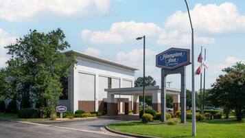 A Hampton Inn by Hilton hotel entrance with a clear sky, an American flag, and a welcoming archway.