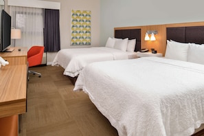 A hotel room with two beds, a desk, a chair, a TV, and a lamp.