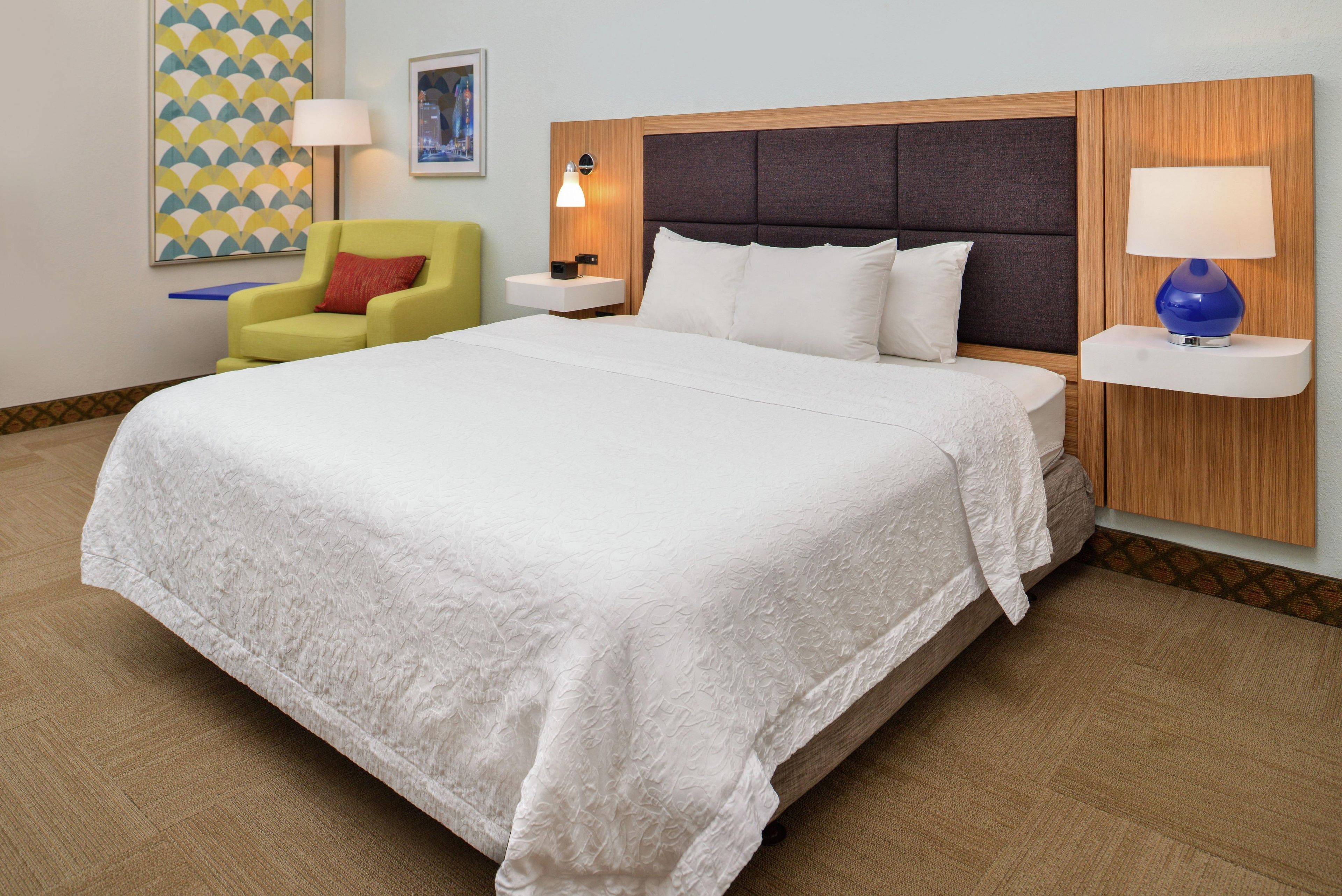 A hotel room with a large bed, a nightstand, a chair, and a wall-mounted lamp.