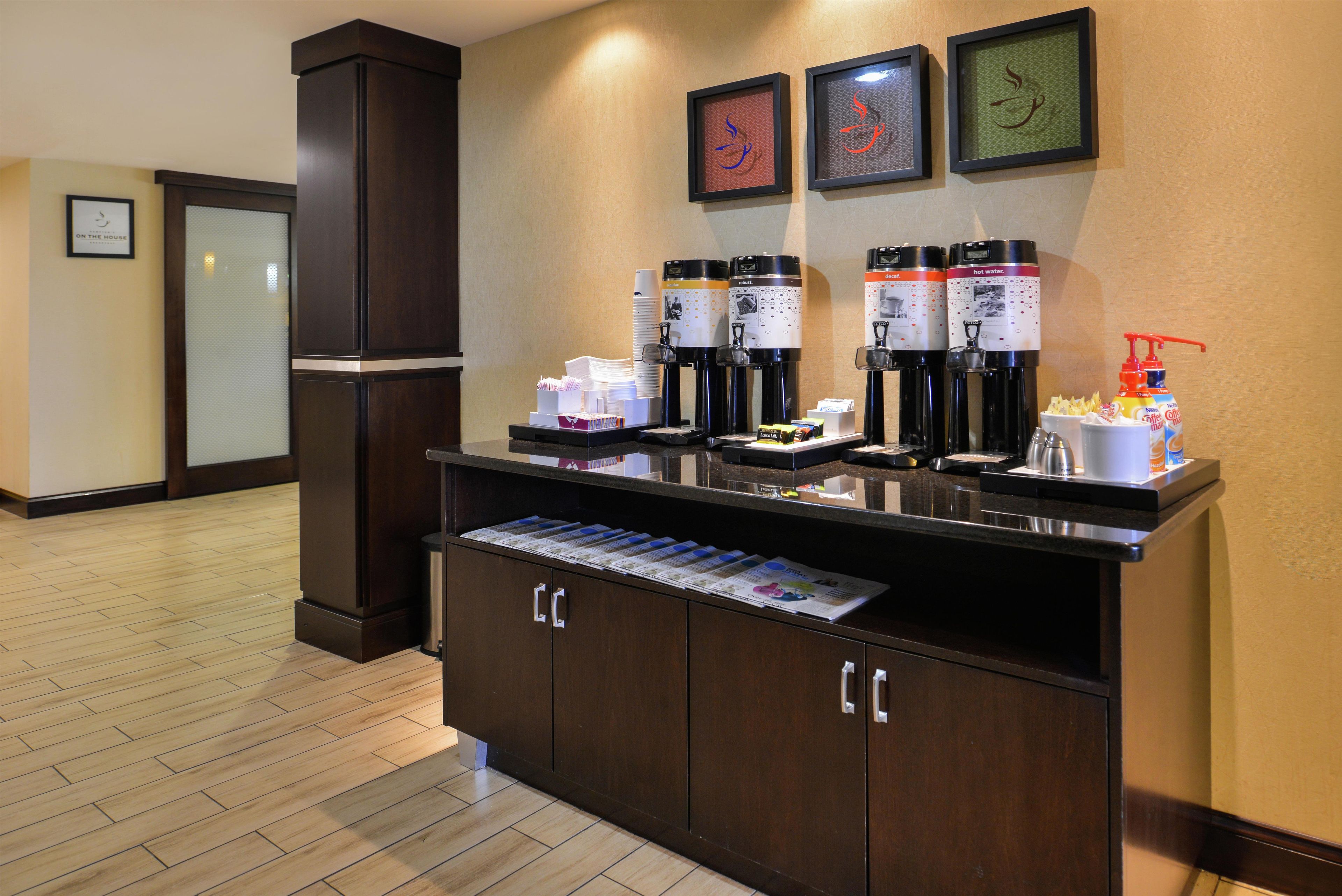 A hotel reception area with a coffee station, framed artwork on the wall, and a magazine rack.