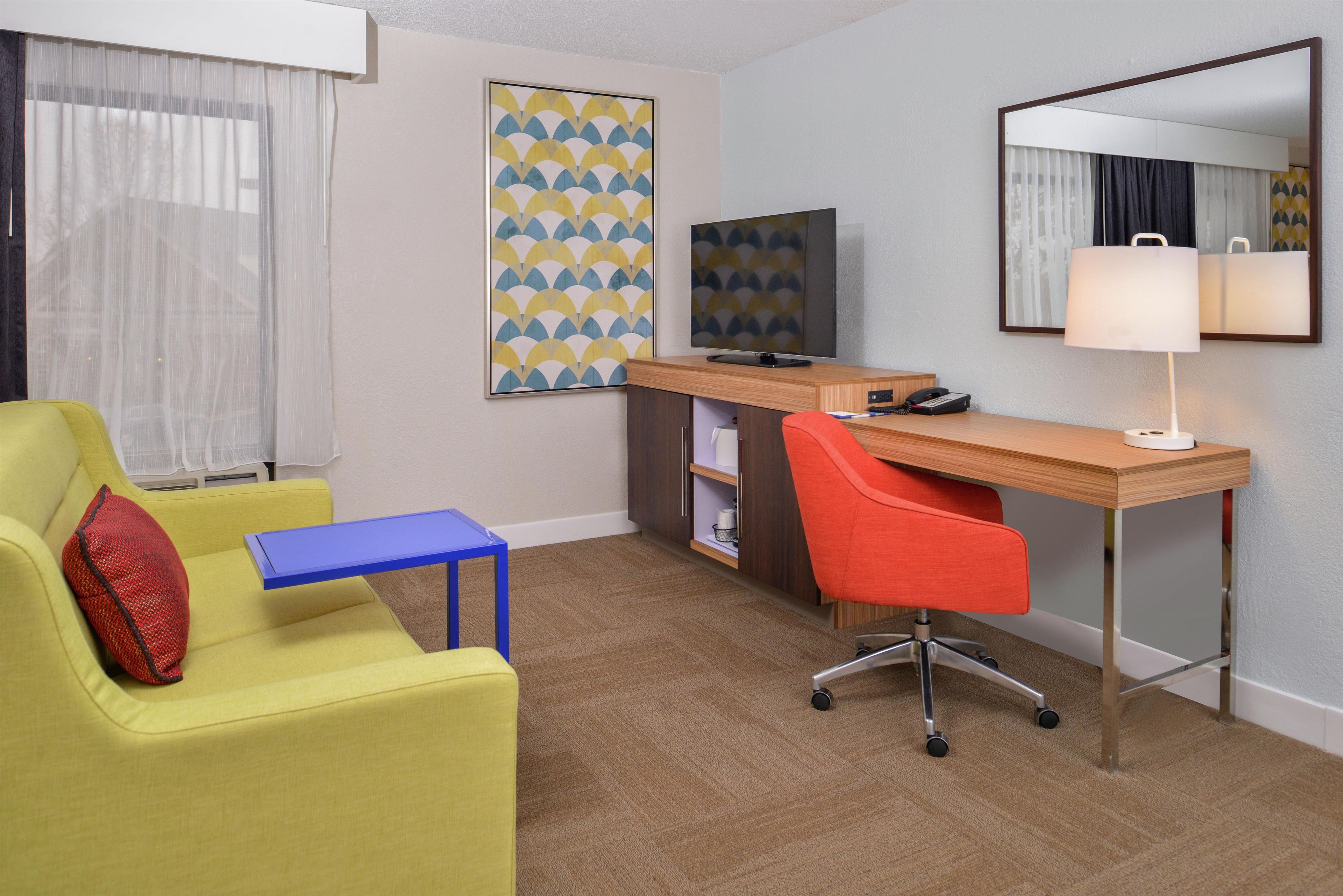 A hotel room with a yellow armchair, a blue side table, a wooden desk with a computer, a red office chair, a television, and a mirror.