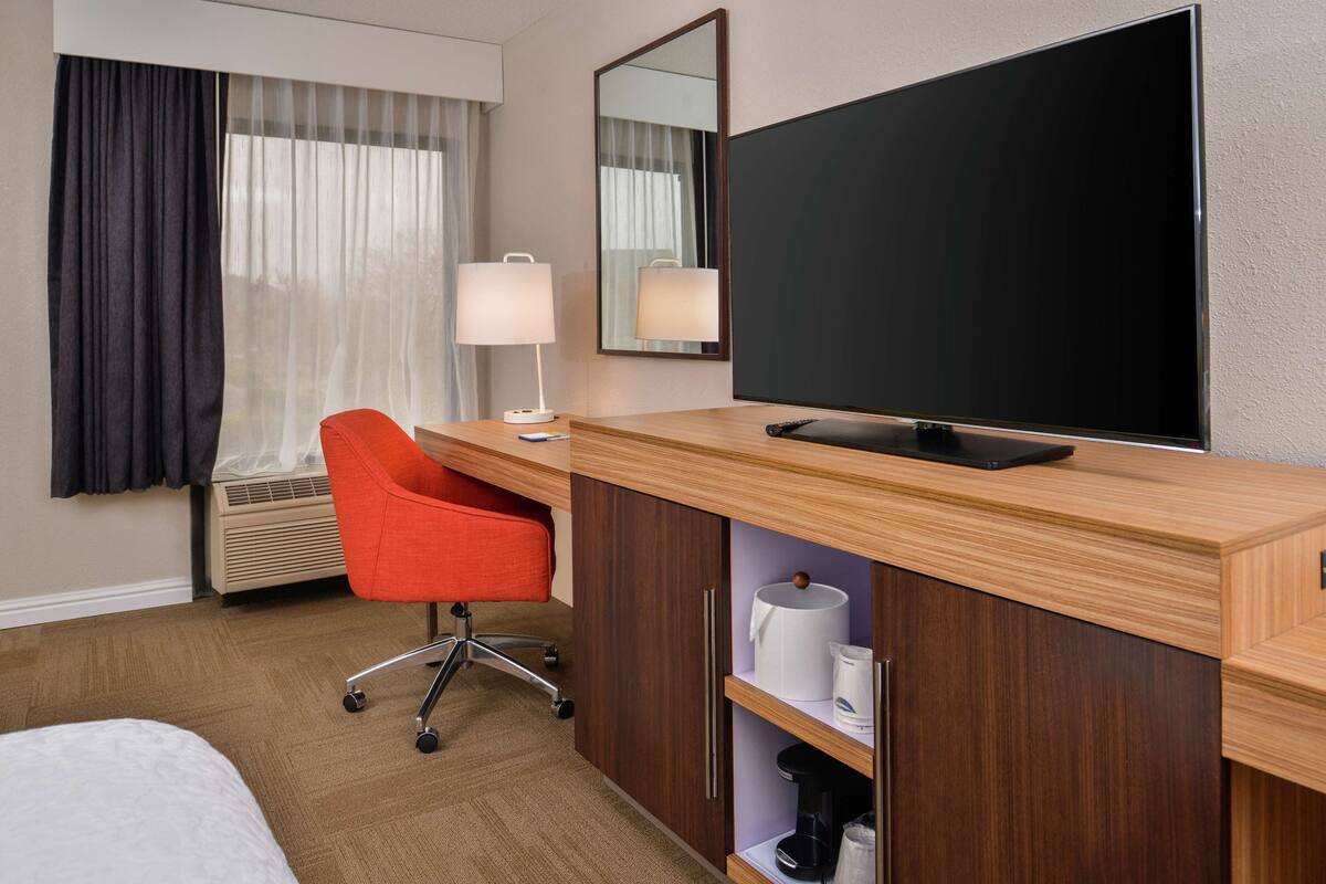 A hotel room with a wooden desk, a red chair, a flat-screen TV, a mirror, and a bed.