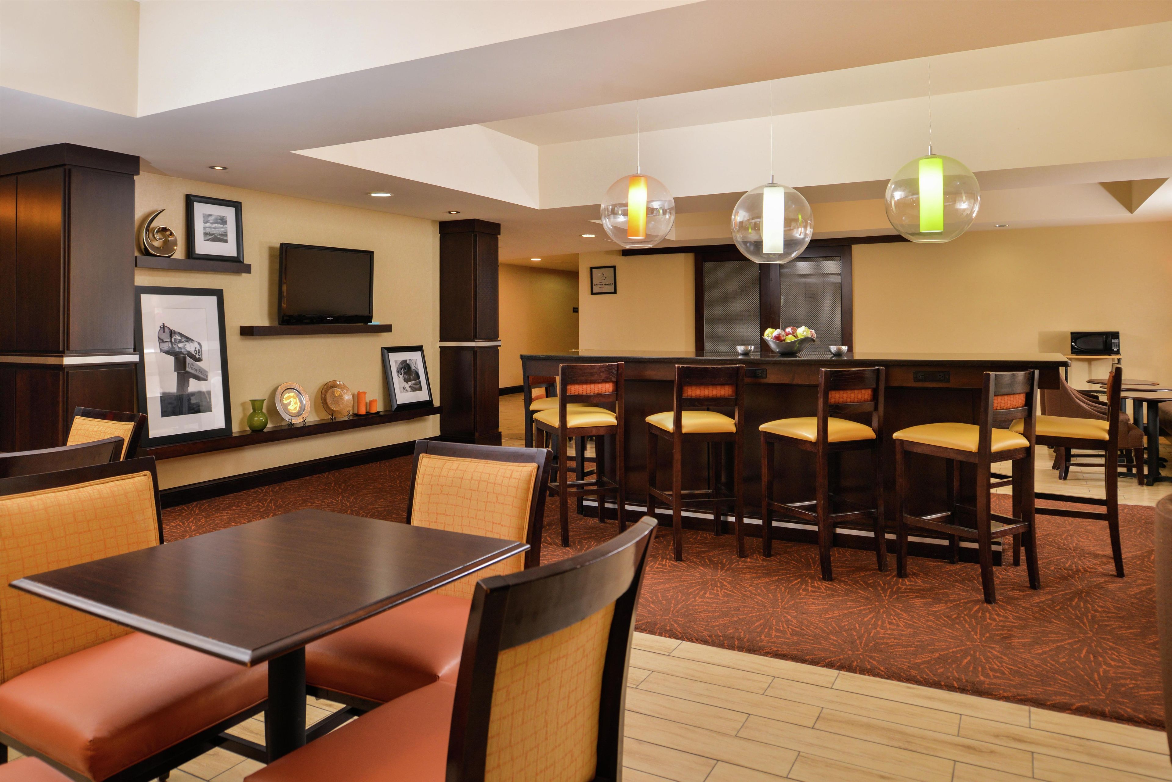 A hotel dining area with multiple tables and chairs, a television on the wall, and pendant lights.