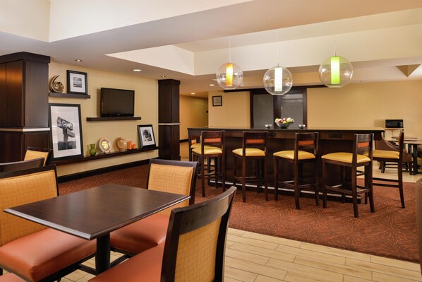A hotel dining area with multiple tables and chairs, a television on the wall, and pendant lights.