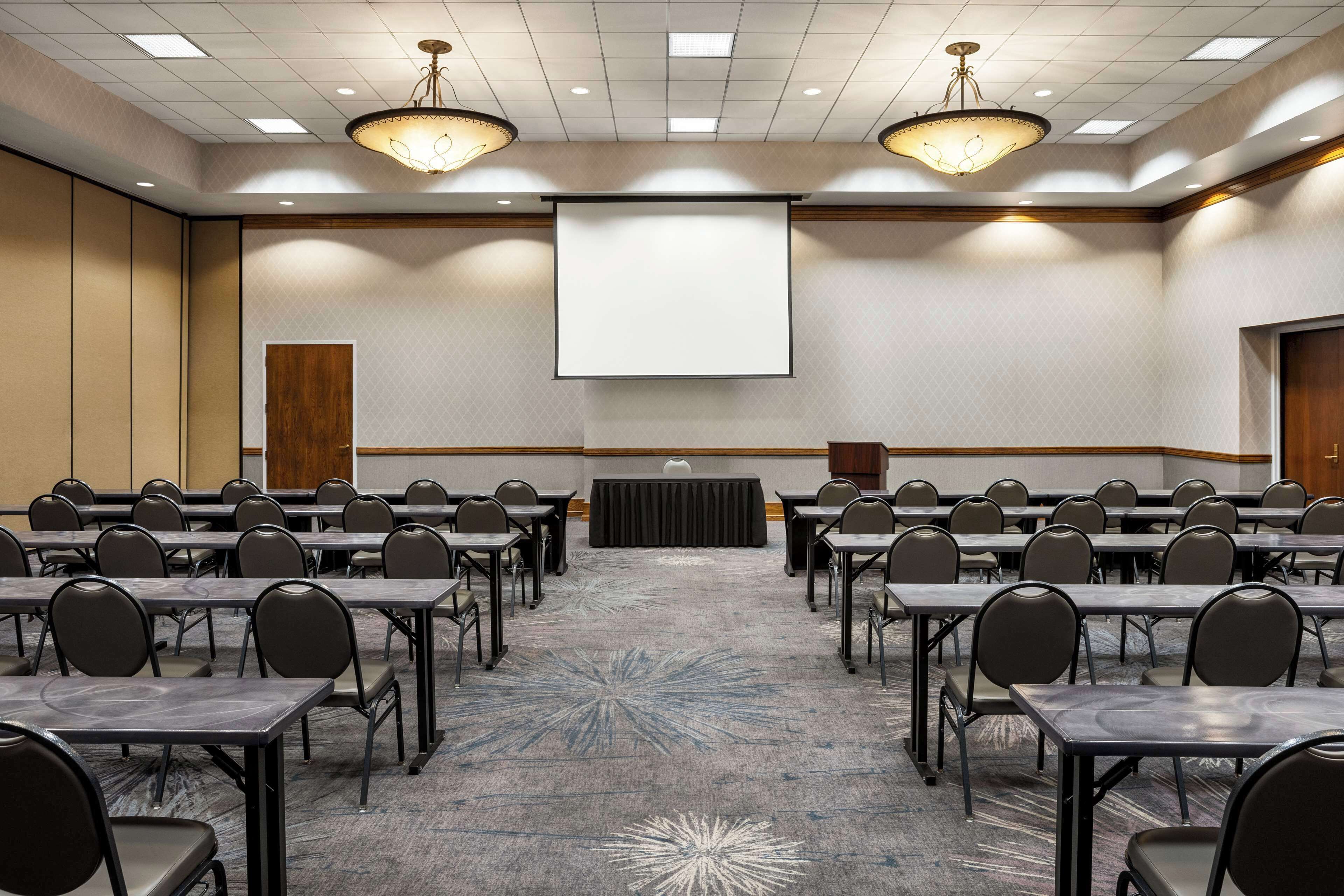 Una sala conferenze con file di tavoli e sedie, uno schermo per proiettore e un pavimento in moquette.