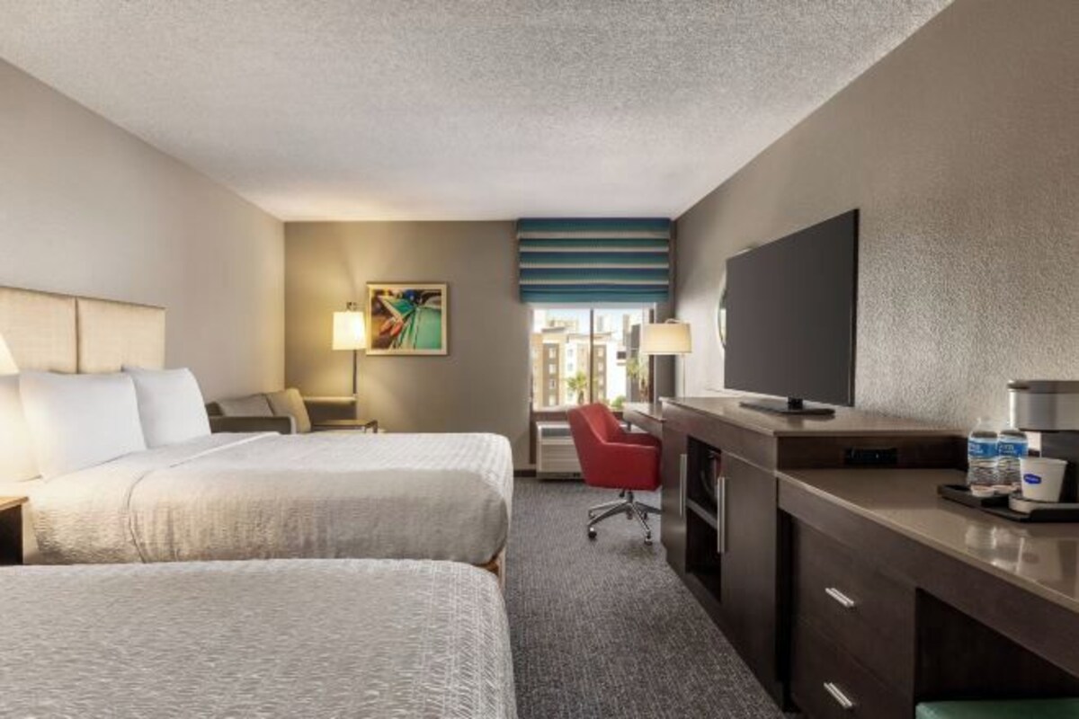 A hotel room with two beds, a desk with a flat-screen TV, a red chair, and a window with a view of buildings.