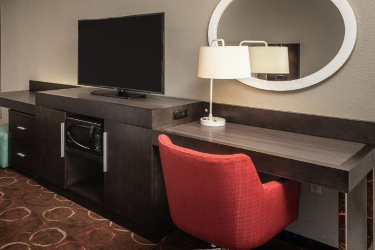A hotel room with a desk, a television, a mirror, and a red chair.
