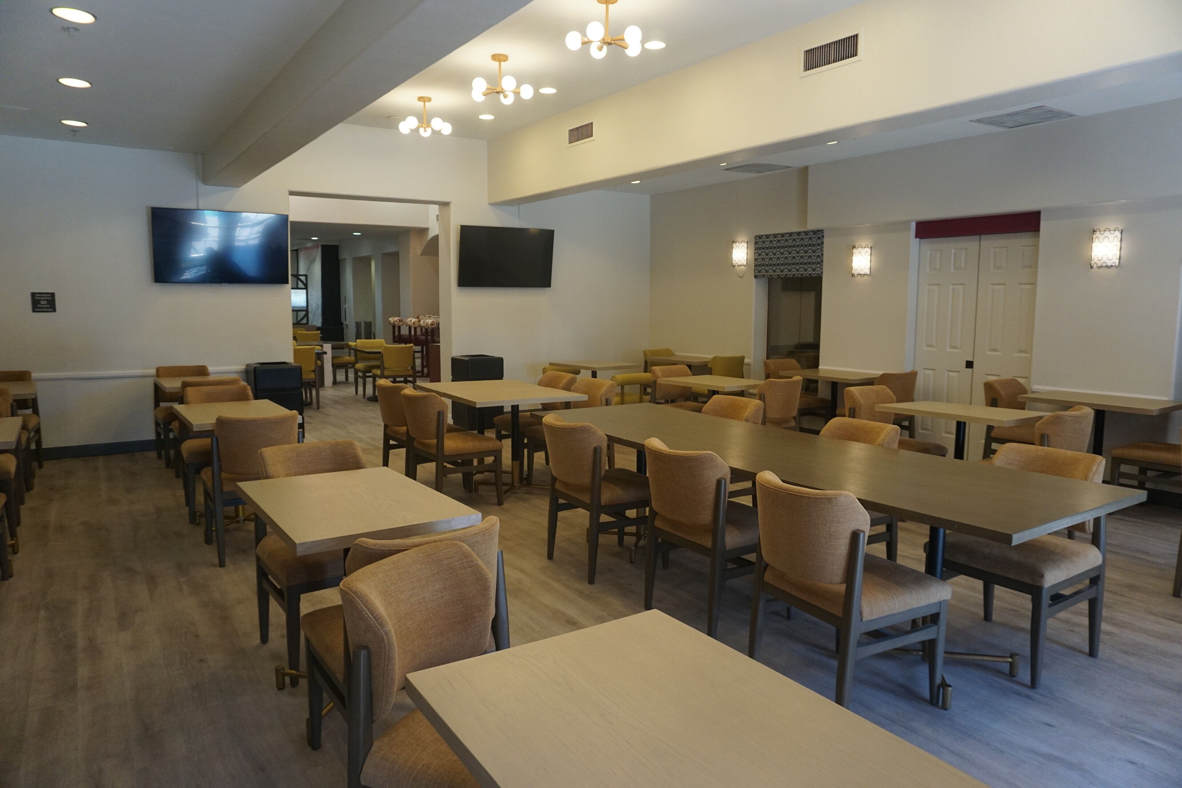 Un grand espace dédié à la salle à manger, avec plusieurs tables et chaises, un téléviseur fixé au mur et une porte dont l’encadrement est de couleur rouge.