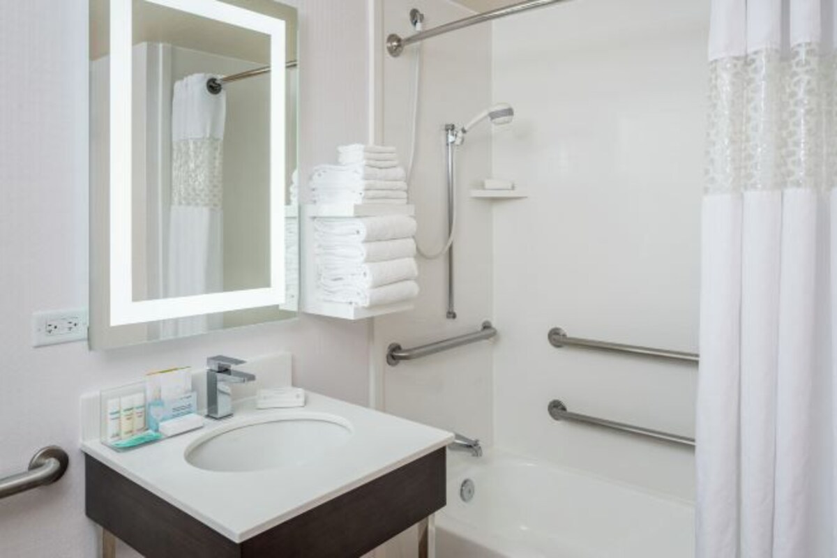 A modern bathroom with a white sink, a mirror, a shower with grab bars, and neatly stacked towels.