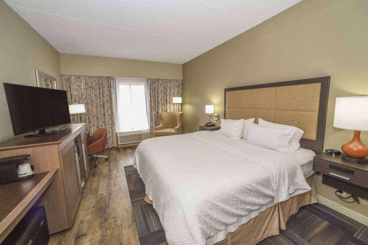 Ein Hotelzimmer mit einem großen Bett, einem Schreibtisch mit Fernseher, einem Sessel und einem Fenster mit Vorhängen.