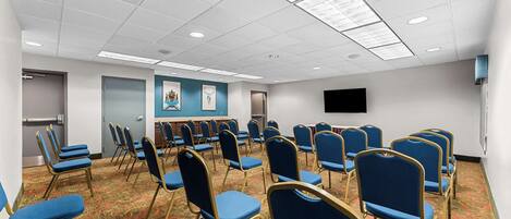A conference room with rows of blue chairs, a carpeted floor, and a flat-screen TV on the wall.