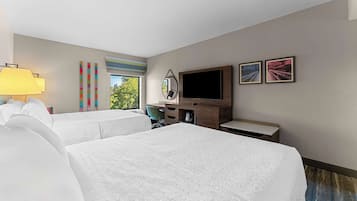 A hotel room with two beds, a desk, a TV, and a window with a view of trees.