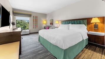 A hotel room with a large bed, a desk, a chair, a TV, and a window with a view of trees.