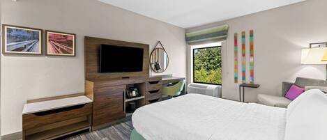 A hotel room with a bed, a TV, a desk, and a window with a view of greenery.