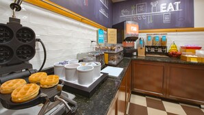Estação de café da manhã com máquinas de waffle, cafeteiras e diversas opções para o café.