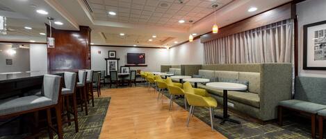 A hotel lobby with wooden flooring, yellow chairs, a green sofa, and a television on the wall.