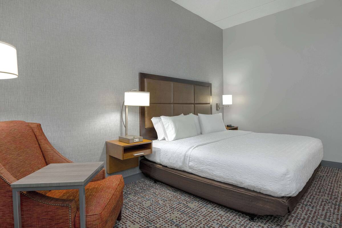A hotel room with a bed, a chair, a nightstand, and a lamp.