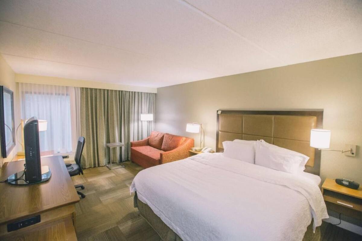 A hotel room with a bed, a desk with a computer, a chair, a lamp, and a nightstand with a phone.