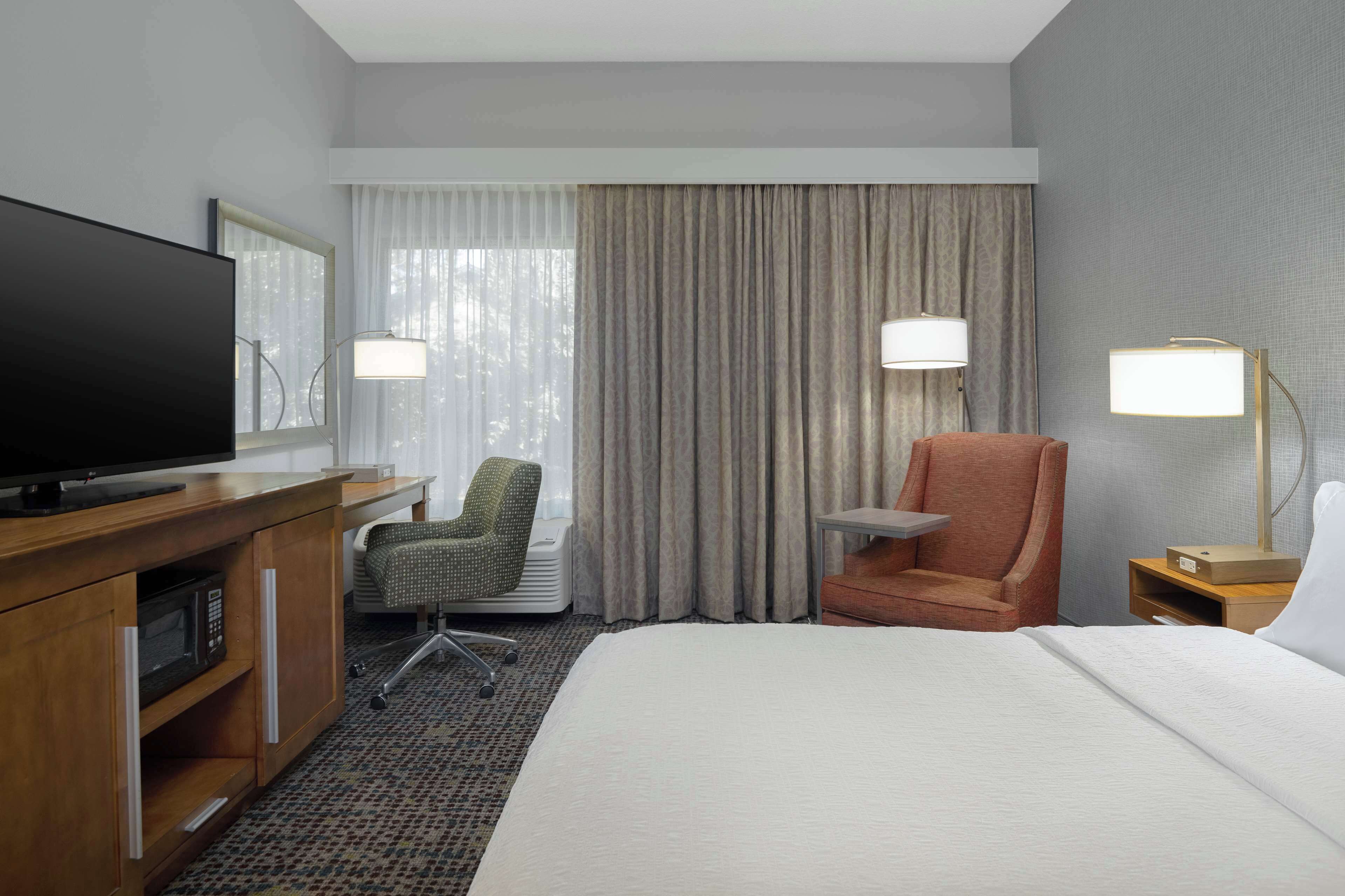 Hotelkamer met een bed, een bureau met televisie, een stoel, een lamp en een raam met gordijnen.