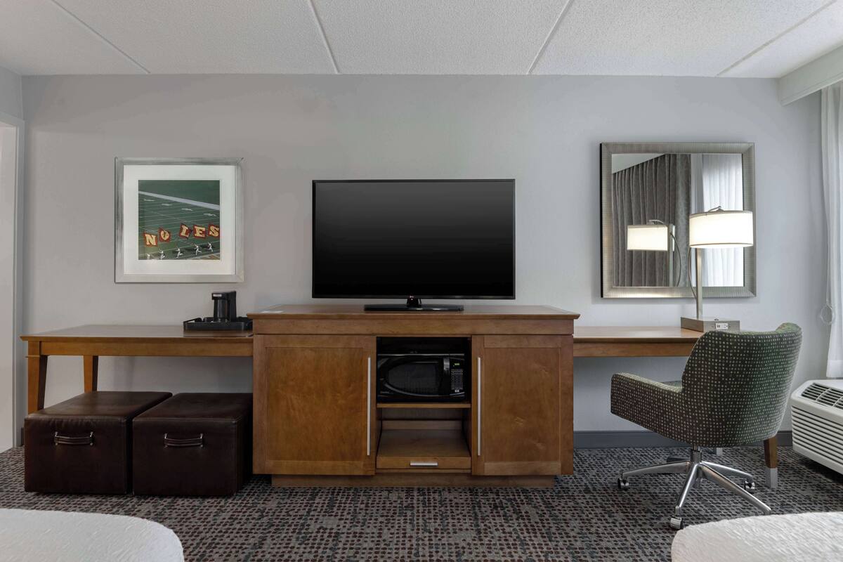 A hotel room with a flat-screen TV, a framed picture, a microwave, a desk, and a chair.
