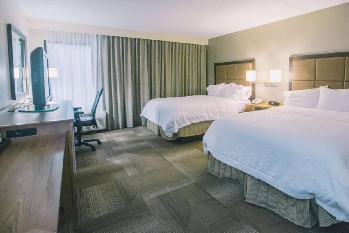 A hotel room with two beds, a desk, a chair, a television, and a window with curtains.