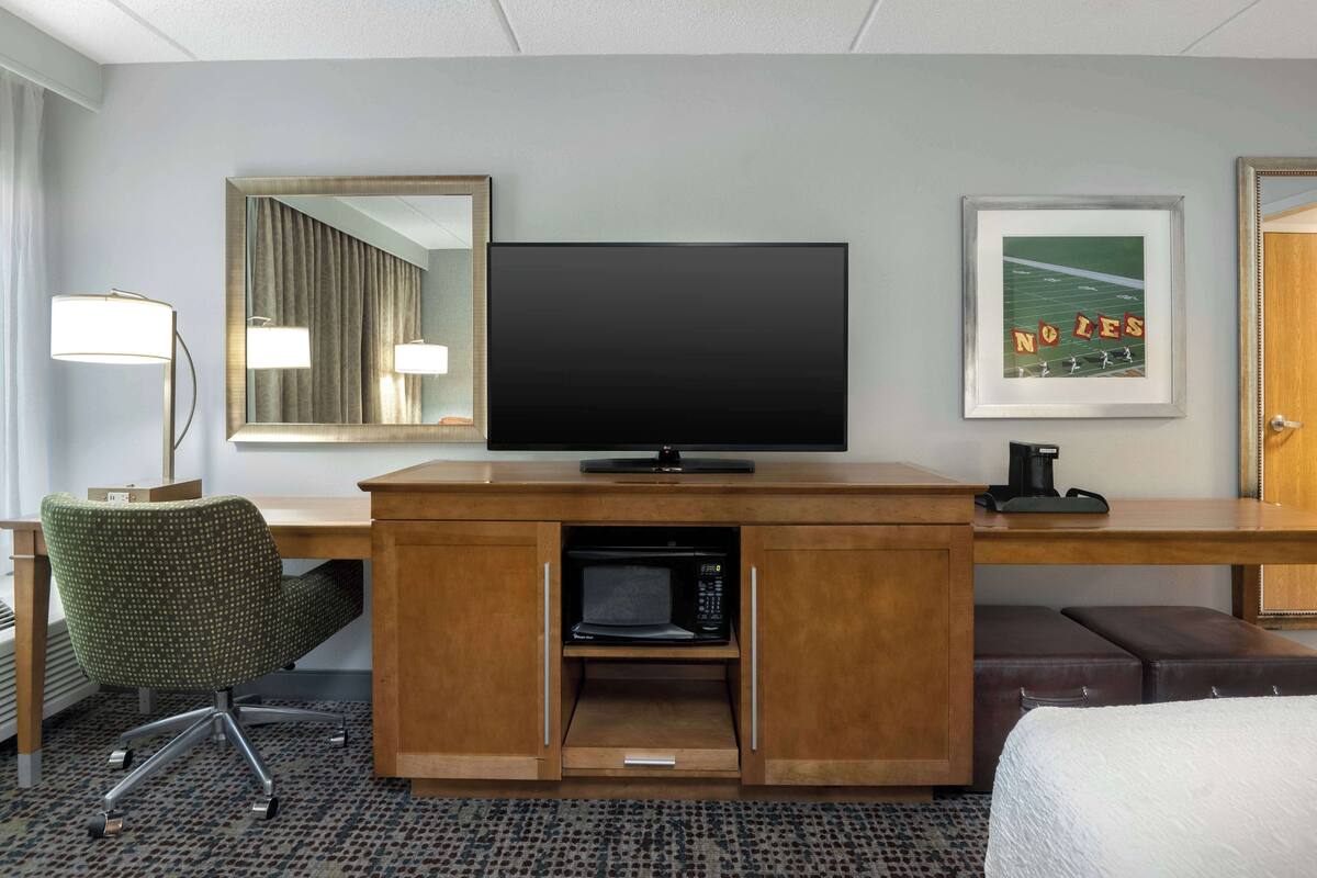 A hotel room with a wooden desk, a flat-screen TV, a microwave, a green office chair, and a framed picture on the wall.