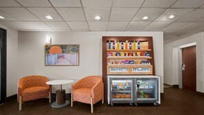 A lobby with two orange armchairs, a small round table, a refrigerator stocked with beverages, and a wall-mounted shelf with snacks. - Hampton Inn Winfield/teays Valley (Hurricane)