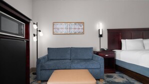 A hotel room with a sofa, a bedside table, a lamp, a bed, and a wall art piece. - Hampton Inn Winfield/teays Valley (Hurricane)