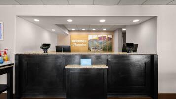 A reception desk with a 'welcome to Hampton' sign, a 'Hilton Honors' plaque, and a digital kiosk.