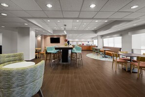 A modern hotel lobby with a central seating area, a reception desk, and a large window providing natural light.
