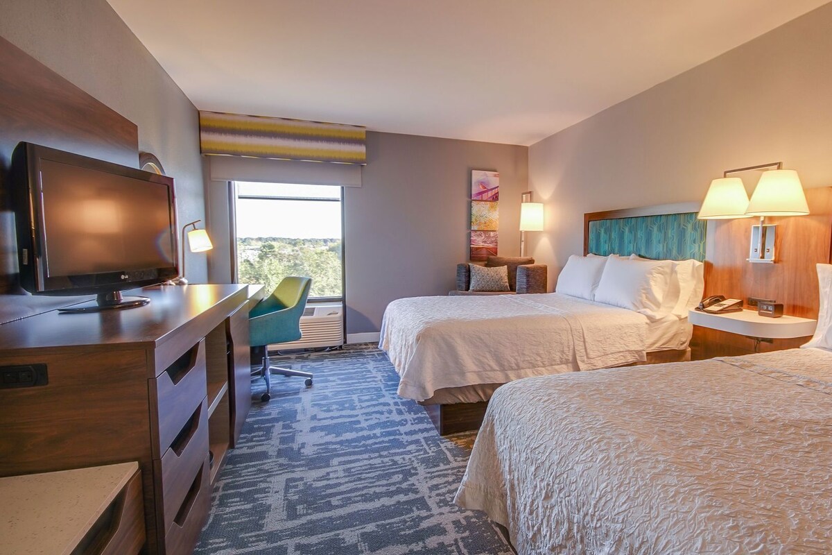 A hotel room with two beds, a desk with a TV, a chair, and a window with a view.