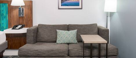 Quarto de hotel moderno com um sofá cinzento, uma pequena mesa de madeira, um quadro com uma vista urbana enquadrada na parede e uma luminária em cada lado do sofá.
