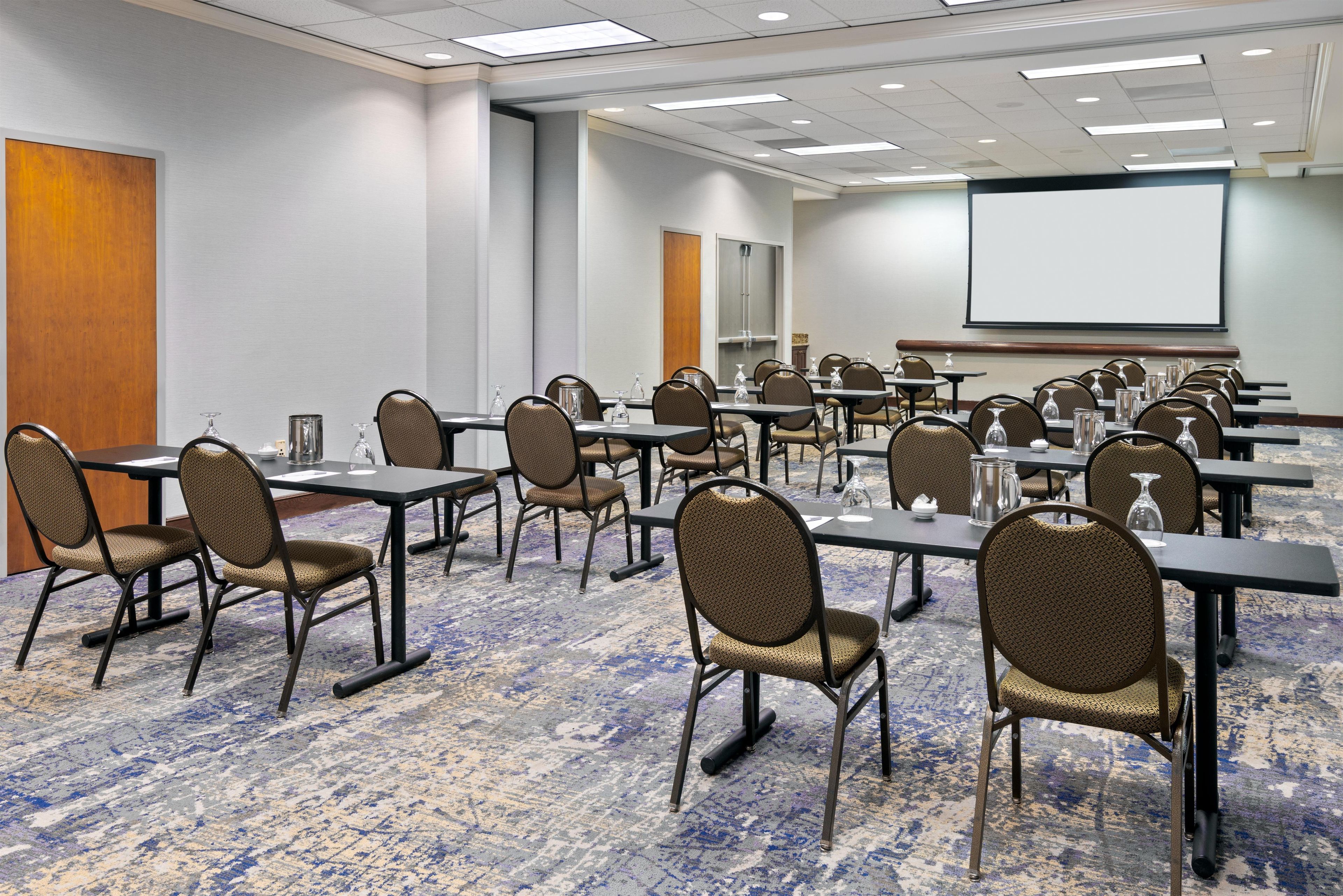 A conference room with rows of tables and chairs, a projector screen, and a carpeted floor.