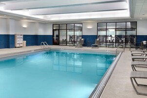 Una piscina interior con agua cristalina, pared azul con azulejos y una zona de estar con sillas y una sombrilla.