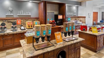 Cocina de hotel con estaciones de autoservicio, que incluyen máquinas para hacer gofres, cafeteras y un cartel que indica necesidades dietéticas.