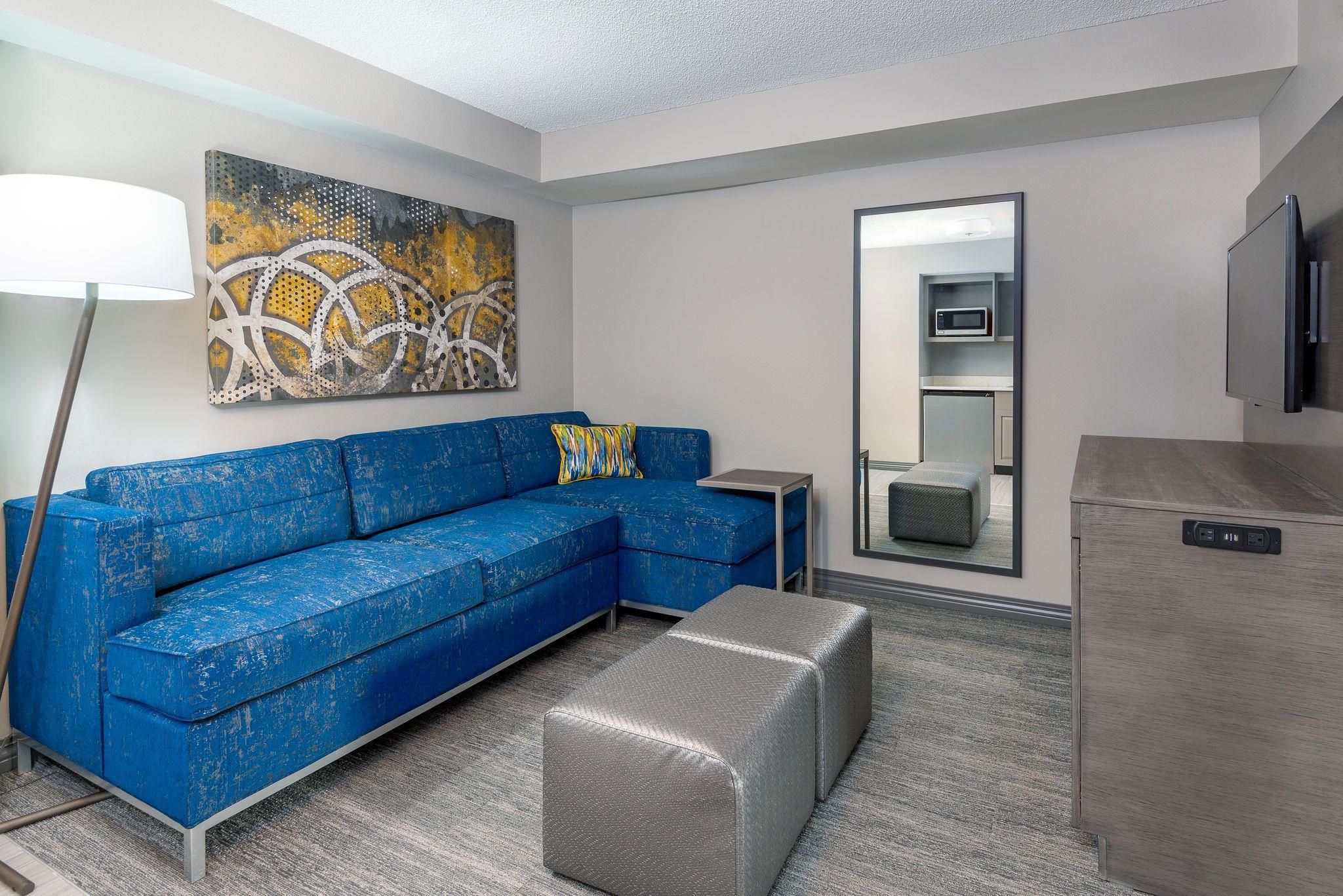 A modern living room with a blue sectional sofa, a grey ottoman, a floor lamp, a painting, a microwave, and a TV.