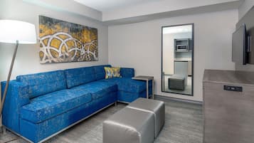 A modern living room with a blue sectional sofa, a grey ottoman, a floor lamp, a painting, a microwave, and a TV.