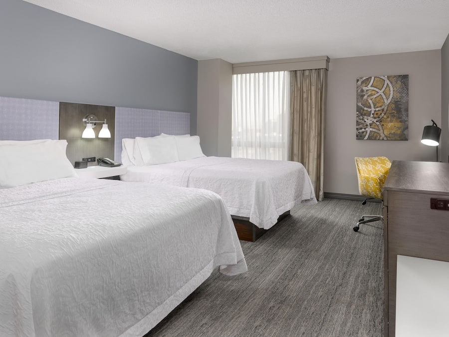 A hotel room with two beds, a desk, a chair, a painting, and a TV.
