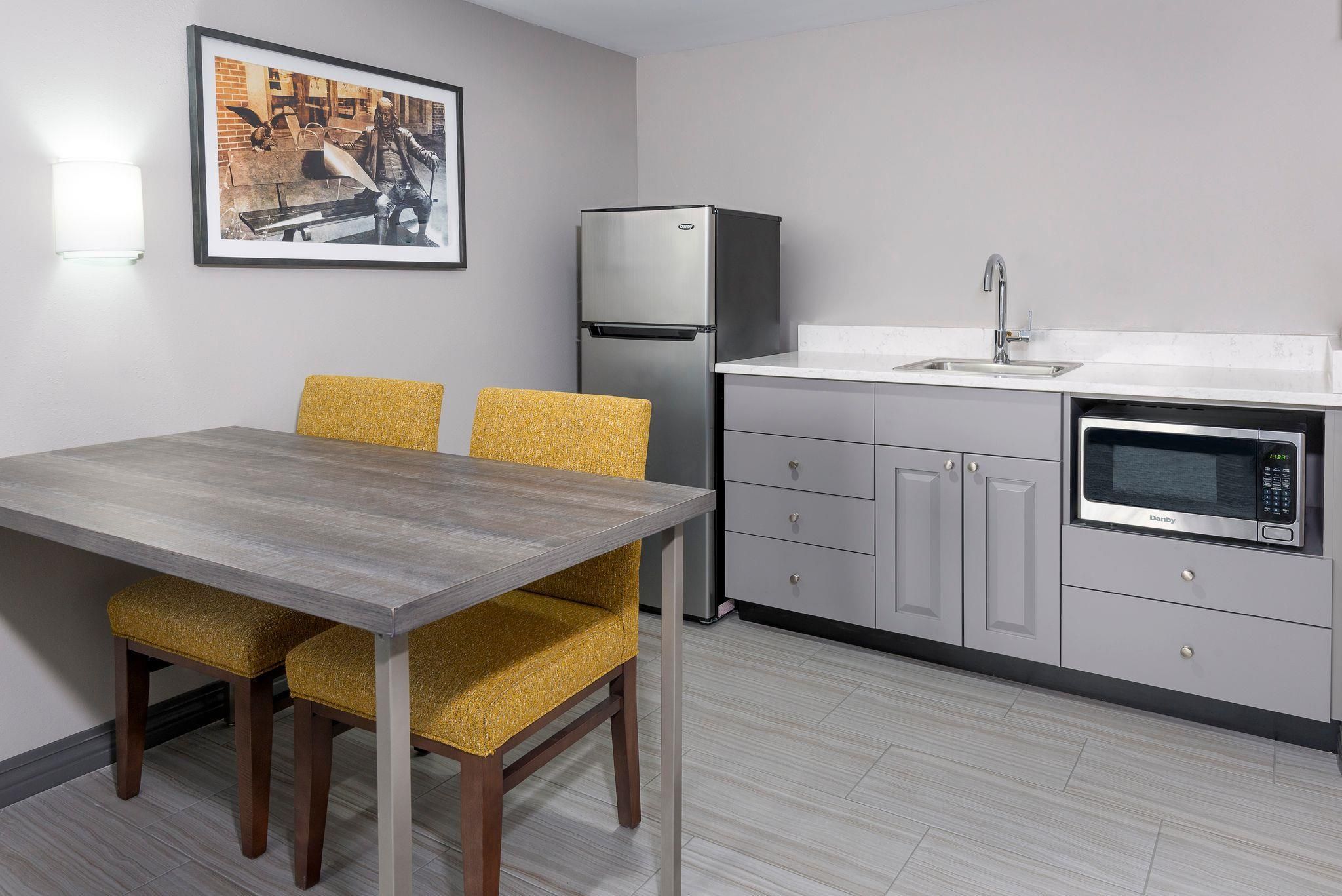 Una cocina moderna con refrigerador, microondas y lavamanos. Hay una mesa de comedor con sillas y un cuadro en la pared.