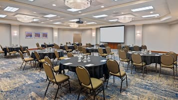 A conference room with multiple tables and chairs, a projector screen, and framed pictures on the wall.